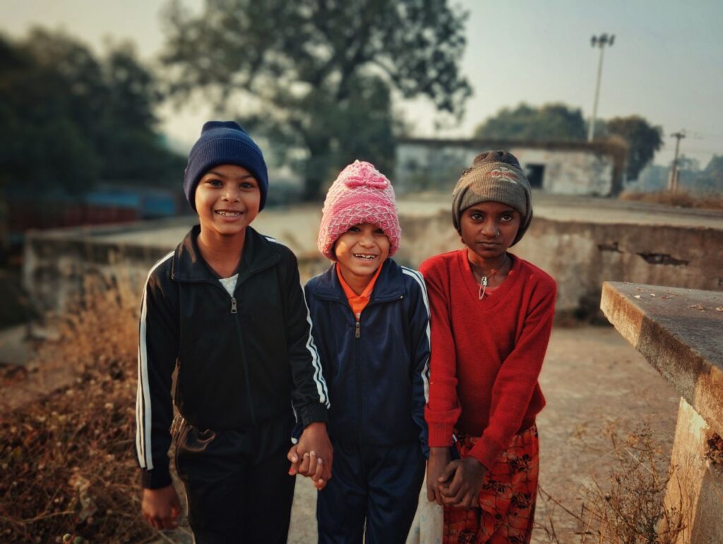 Three cheerful children wearing winter clothes stand together holding hands in a rural village.